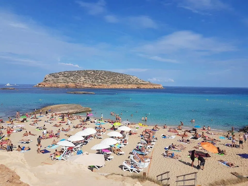 Cala beach in Ibiza with turquoise water, sunbathers, and a rocky island view.
