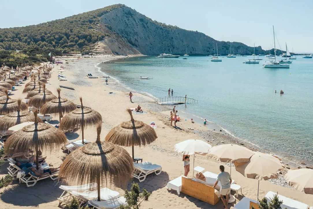 Ibiza beach with sun umbrellas, clear water, boats, and rocky hills.