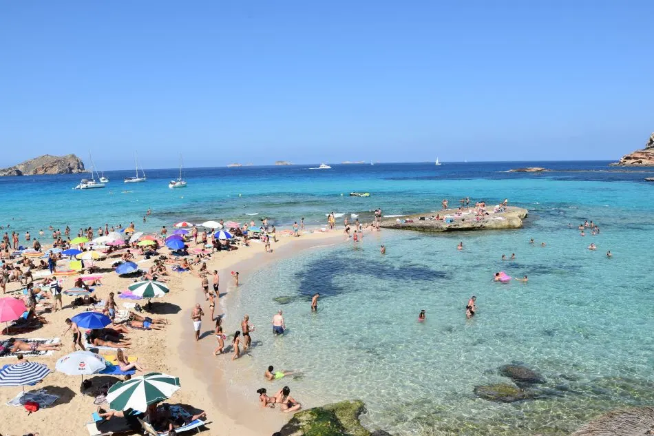 Northern Ibiza beach with crystal-clear water, sandy shore, umbrellas, and swimmers.